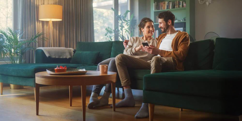 Małżeństwo siedzące na zielonej kanapie w mieszkaniu ze smartfonem w ręce.