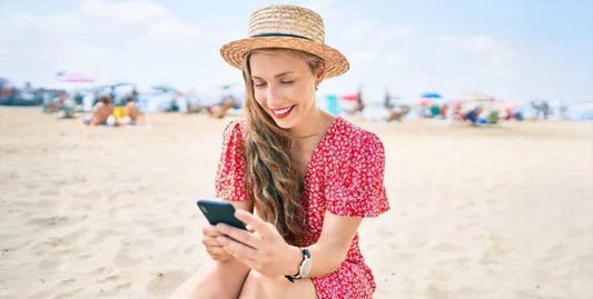 Dziewczyna siedząca na plaży w czerwonej sukience i kapeluszu z telefonem w ręce.