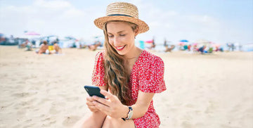 Dziewczyna siedząca na plaży w czerwonej sukience i kapeluszu z telefonem w ręce.