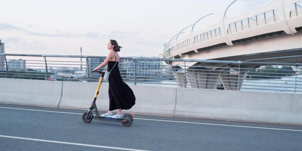 Dziewczyna w czarnej sukience jadąca na hulajnodze elektrycznej po ulicy.