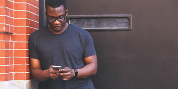 Czarnoskóry mężczyzna szukający numerów telefonów na smartfonie ubrany w czarną koszulkę oparty o ścianę.