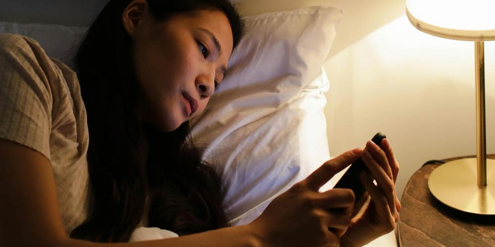 Młoda kobieta przeglądająca smartfon przed snem przy zaświeconej lampce.
