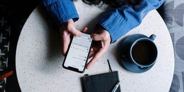 Przeglądanie smartfona przy stole. Na stole kubek z kawą i notes.